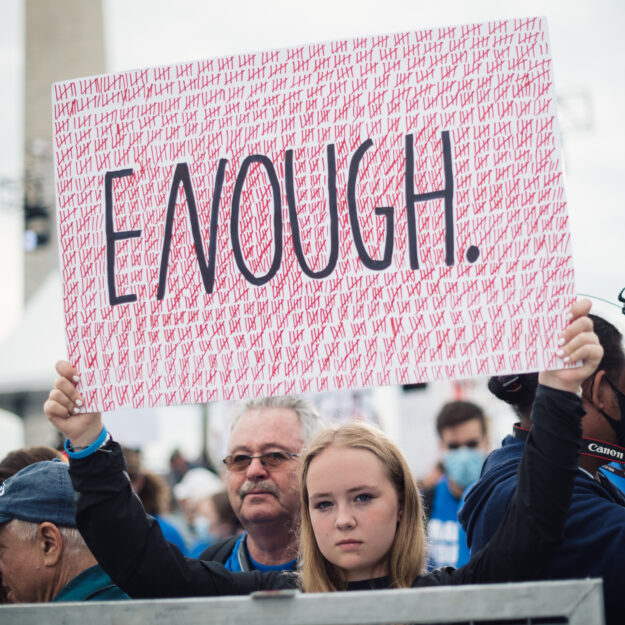 Youth activist holds protest sign reading, "Enough," with hundreds of tally marks to represent lives lost to gun violence.